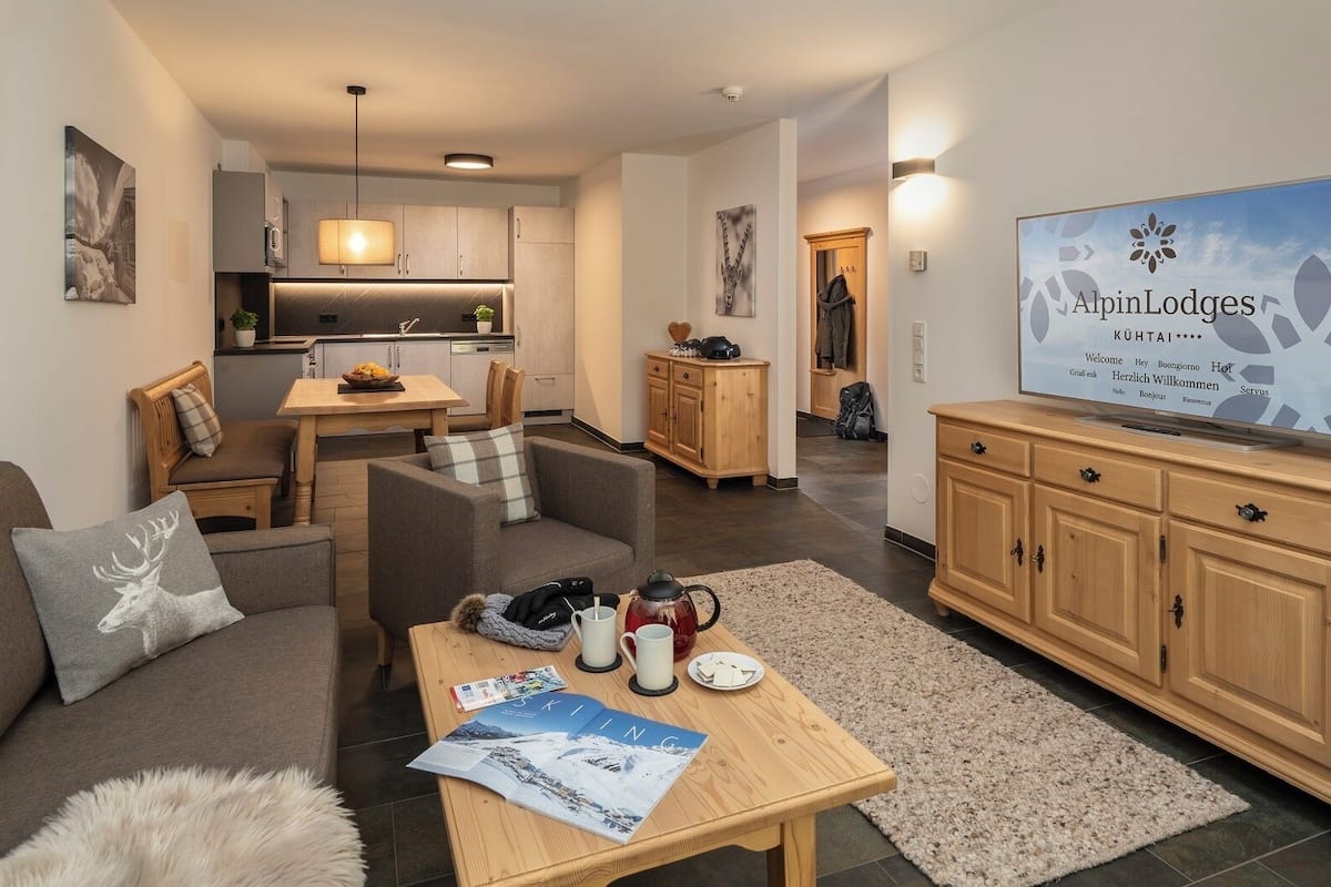 Una sala de estar moderna con sofá, mesa de centro y televisor mostrando el mensaje de bienvenida de 'AlpinLodges'.