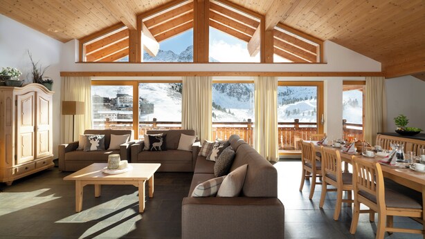 A spacious living area with a high ceiling, wooden beams, and large windows offering mountain views. - AlpinLodges Kühtai (Silz)