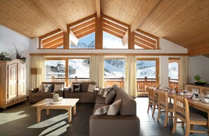 Un vaste espace de vie doté d’un plafond haut, de poutres en bois et de grandes fenêtres offrant une vue sur les montagnes.