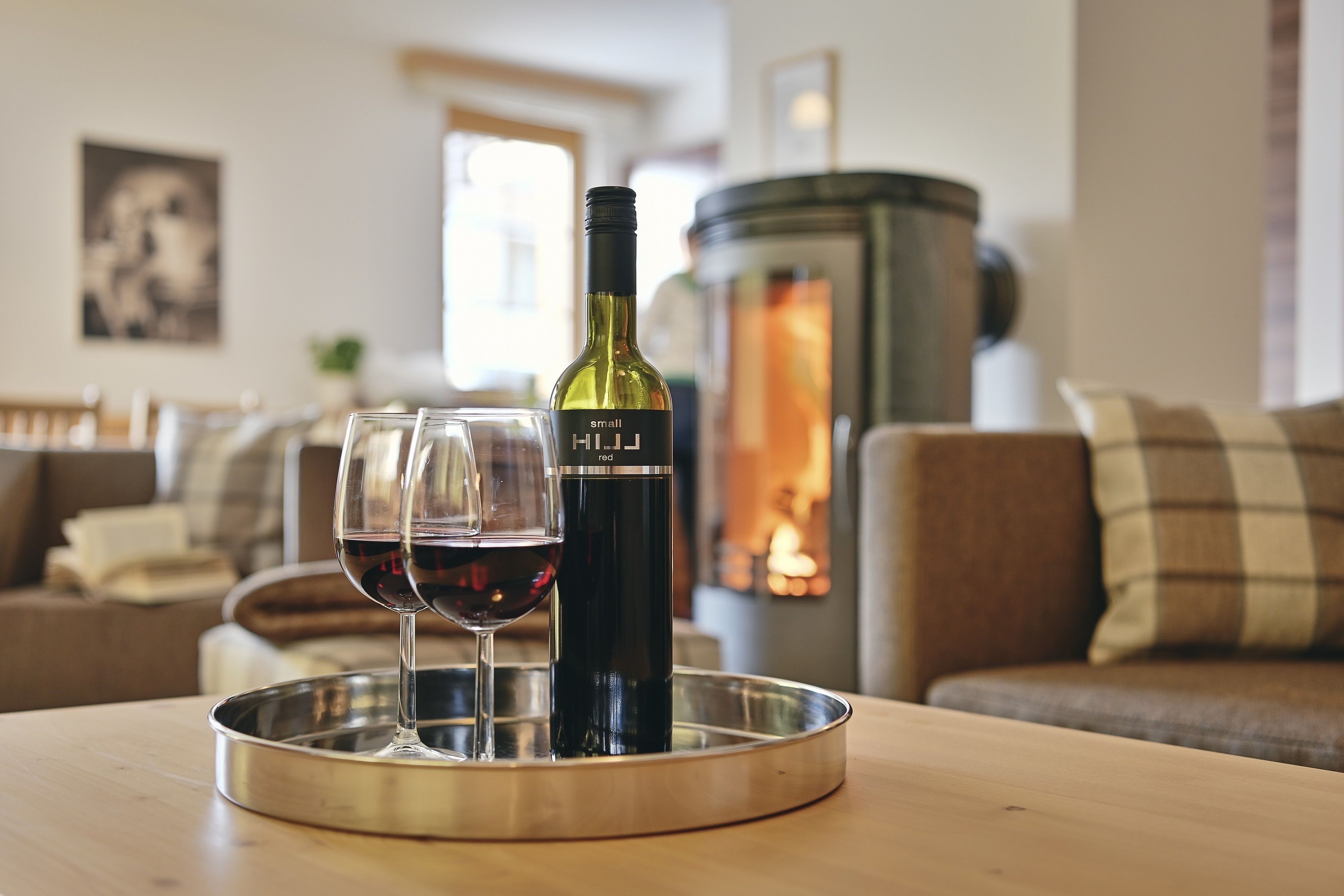 Une bouteille de vin et deux verres sur un plateau, dans un salon chaleureux avec une cheminée.