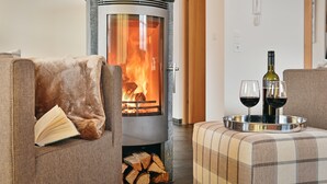 A cozy living room with a wood-burning stove, a book on a chair, and two glasses of red wine on a tray. - AlpinLodges Kühtai (Silz)