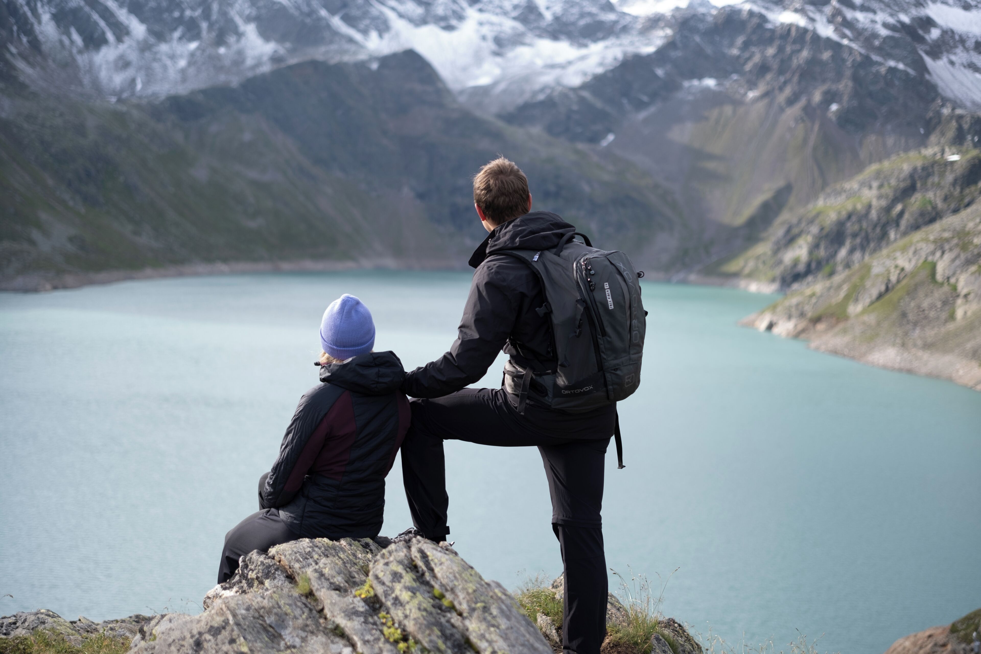 Two people sitting on a rock by a lake with mountains in the background.