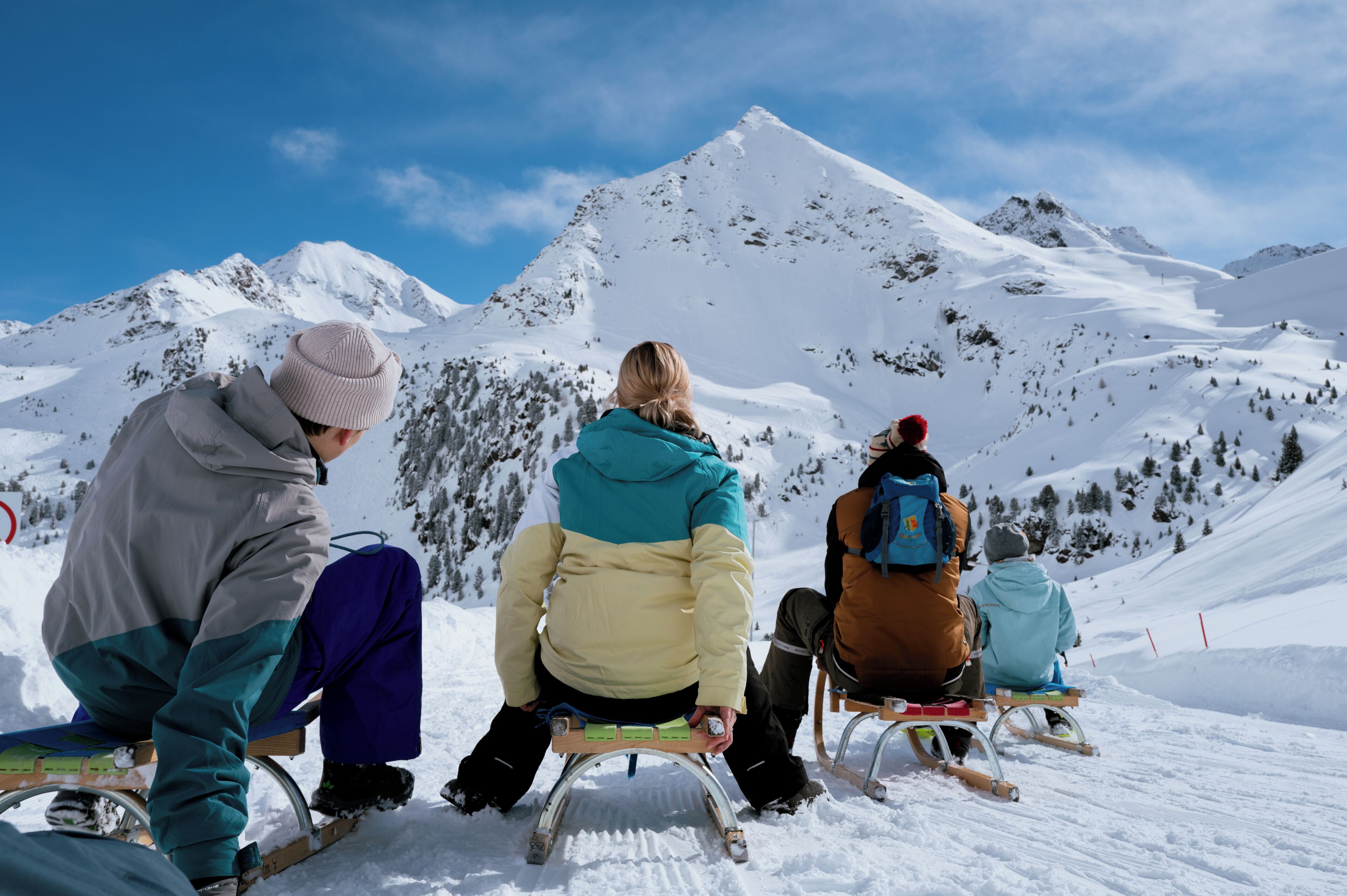 Three people seated on sleds on a snowy slope with mountains in the background.