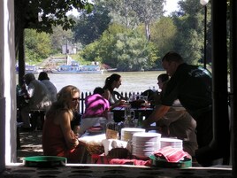 A riverside dining area with tables and chairs, a view of the river, and boats in the background.