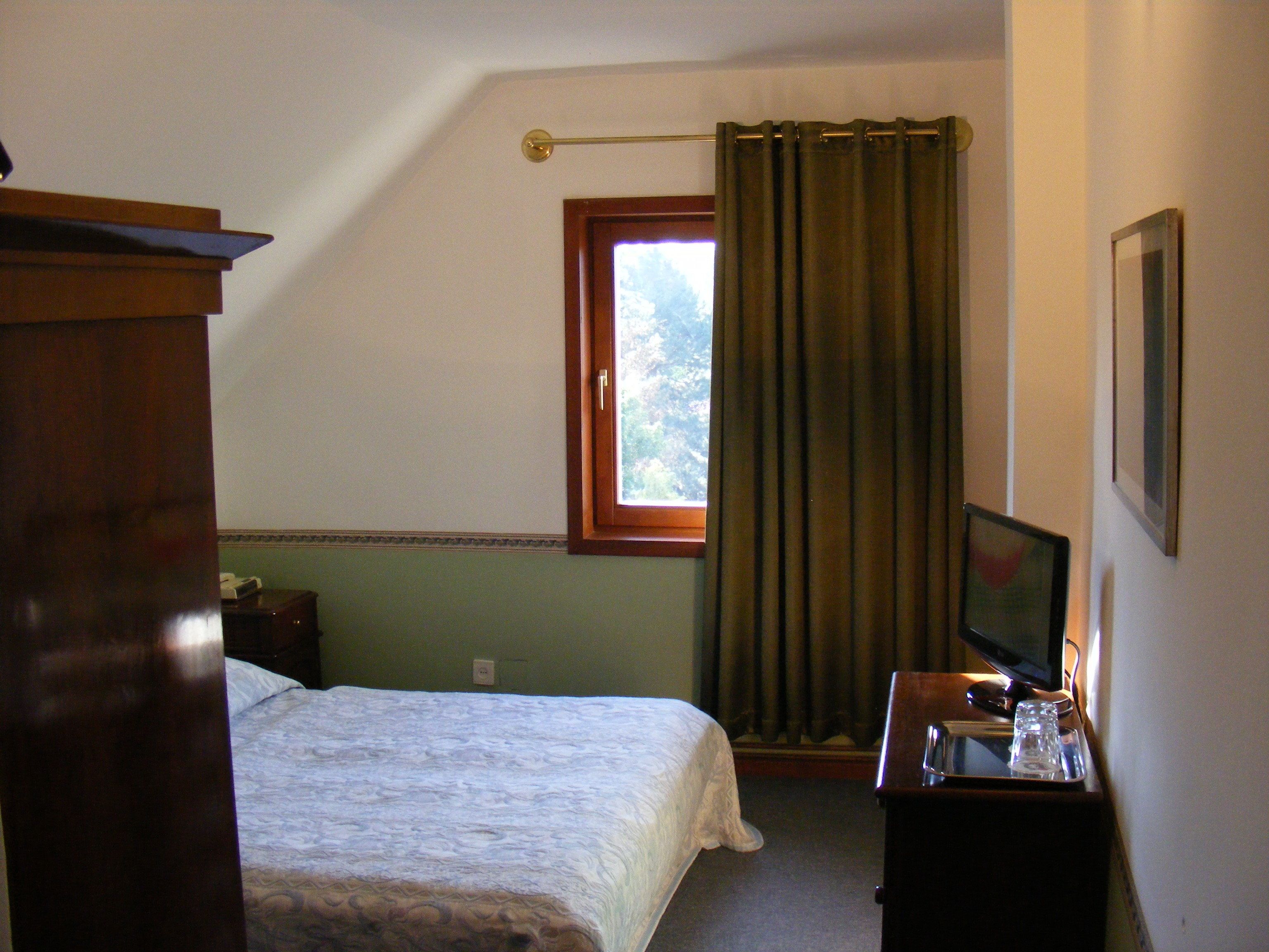 A bedroom with a bed, a desk with a computer, a window with curtains, and a framed picture on the wall.