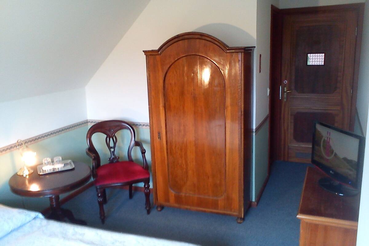 A hotel room with a wooden wardrobe, a wooden chair with a red cushion, a small table with a lamp, and a television on a wooden stand.