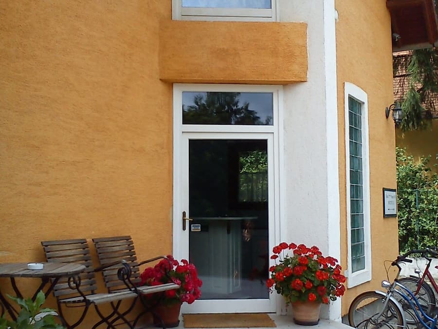 A building with a yellow exterior, a white door, and a small porch with two wooden chairs and a table. There are potted red flowers on the porch and a bicycle parked outside.