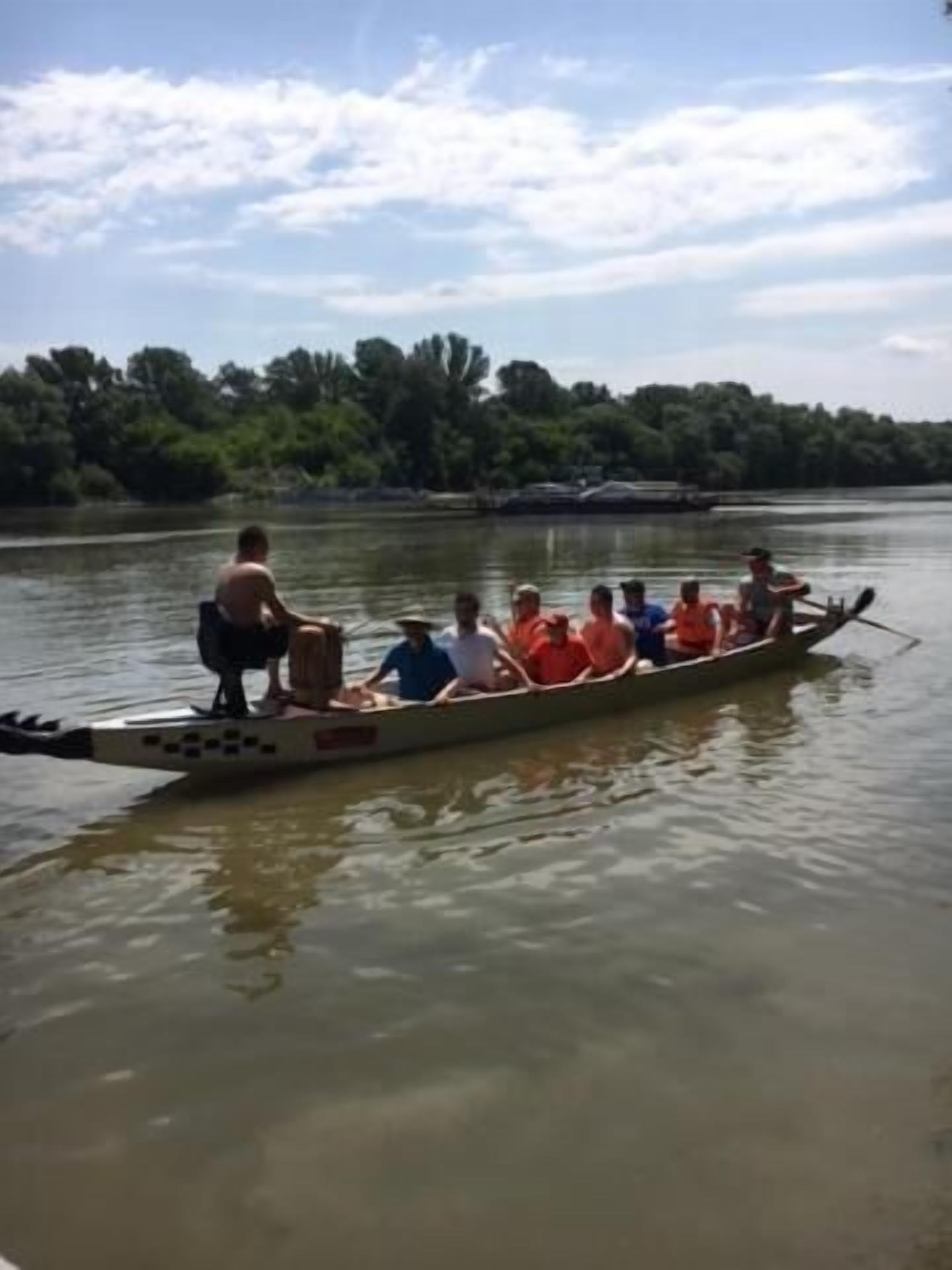 Um barco comprido e estreito com várias pessoas remando, cercado por águas calmas e uma margem coberta por floresta.