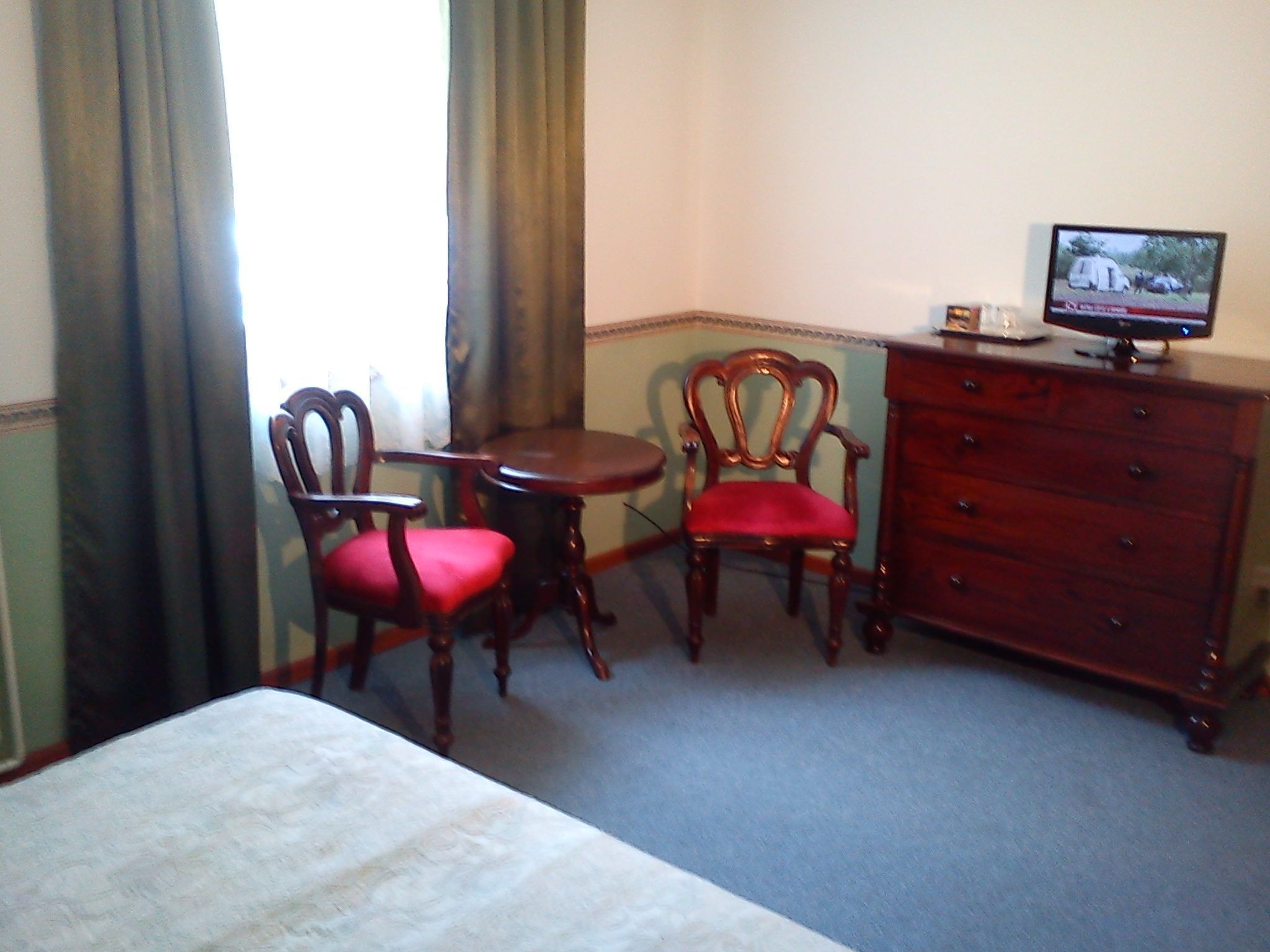 A hotel room with a bed, two wooden chairs with red cushions, a small round table, a wooden dresser, and a television on top of the dresser.