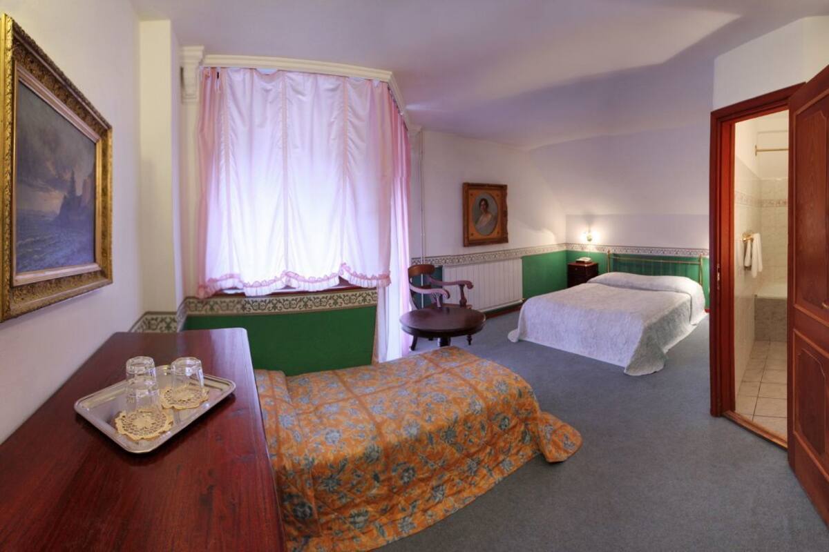 A hotel room with two beds, a wooden desk, a window with curtains, a framed painting, and a portrait on the wall.