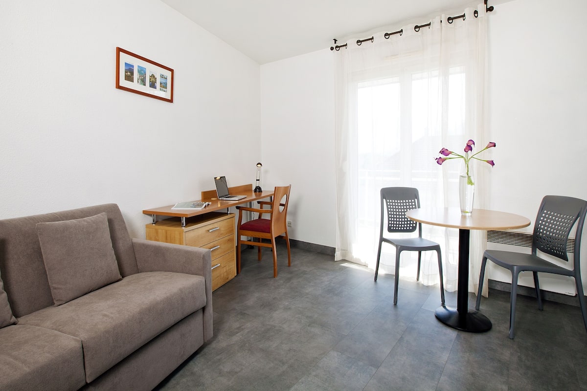 A modern living room with a sofa, a dining table with chairs, a small wooden desk, and a vase with flowers.