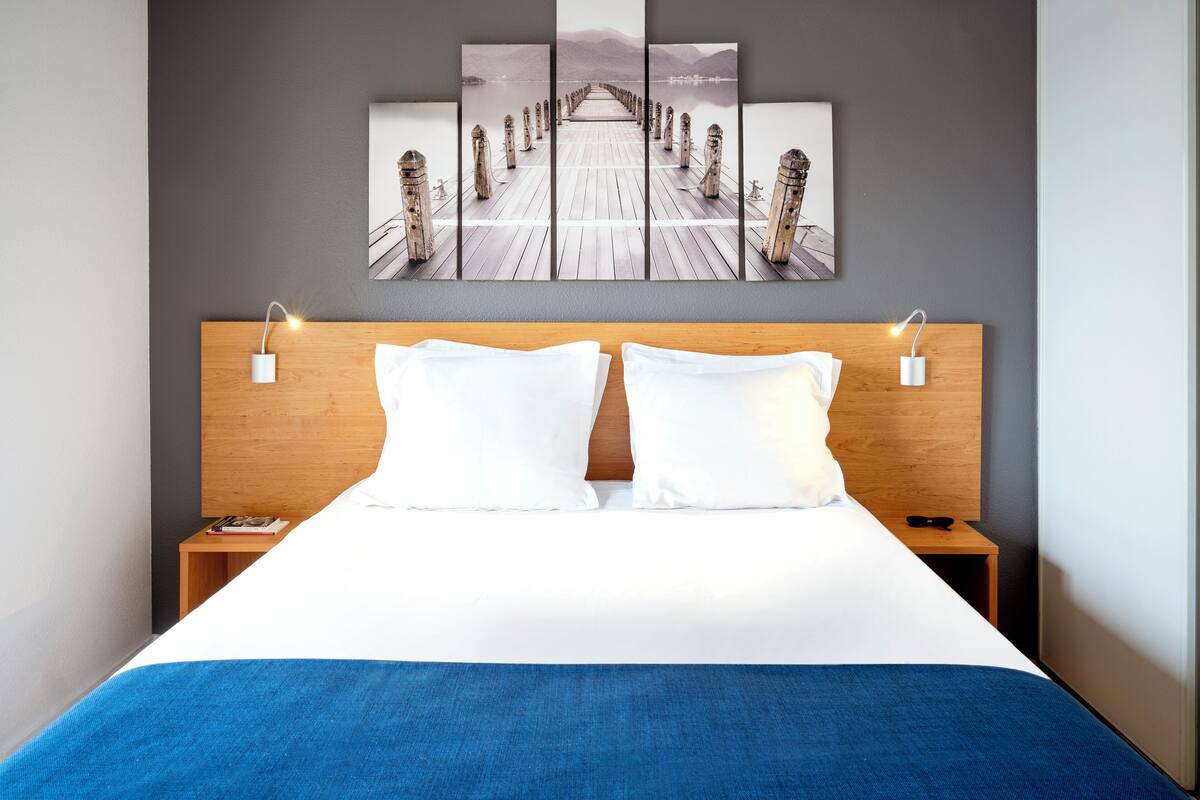A hotel room with a wooden headboard, white bedding, a blue throw blanket, and a large wall art piece depicting a pier.