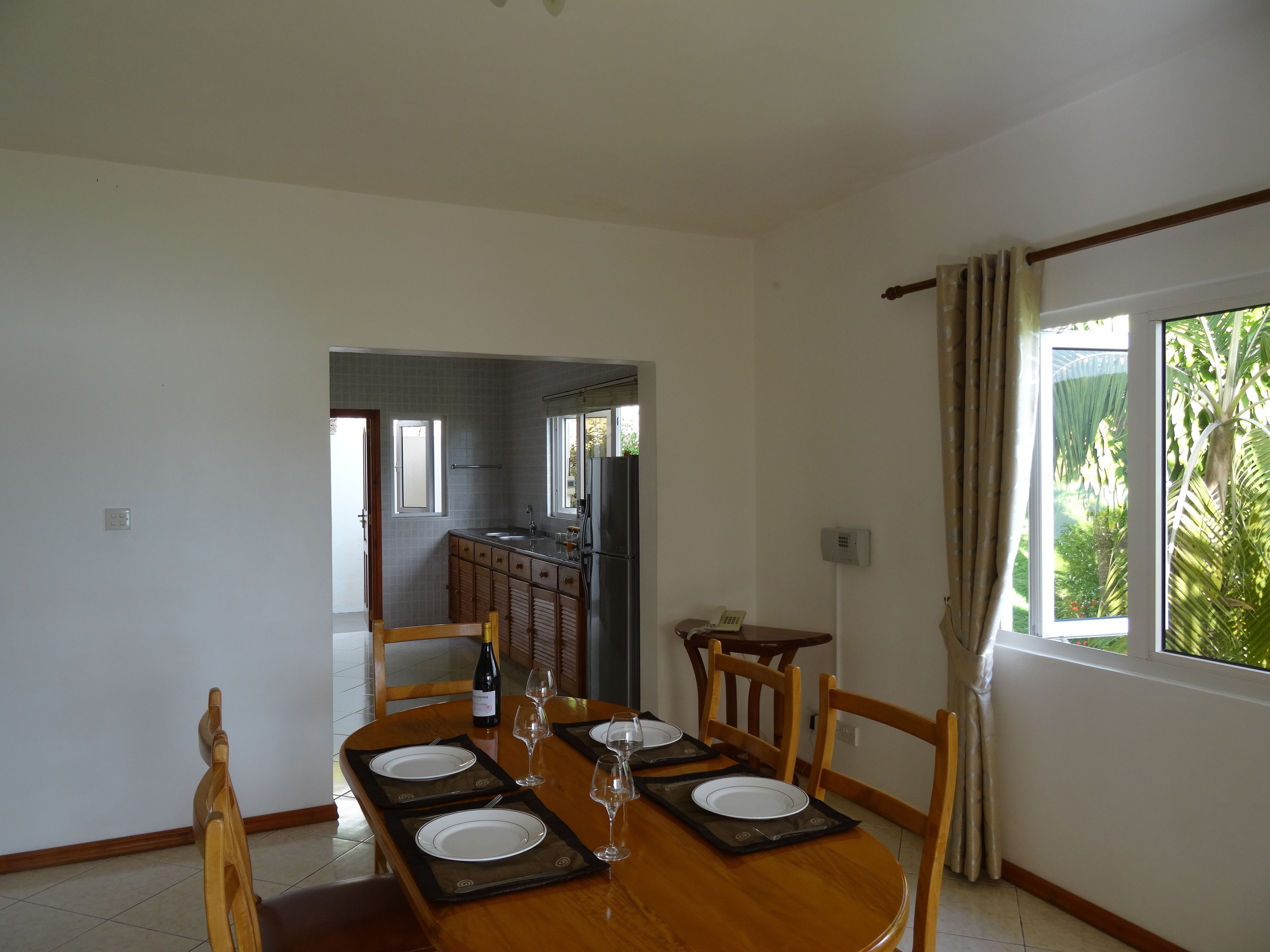 A dining area with a wooden table set for four, a kitchen in the background, and a window with curtains.