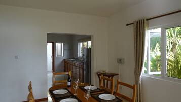 A dining area with a wooden table set for four, a kitchen in the background, and a window with curtains.