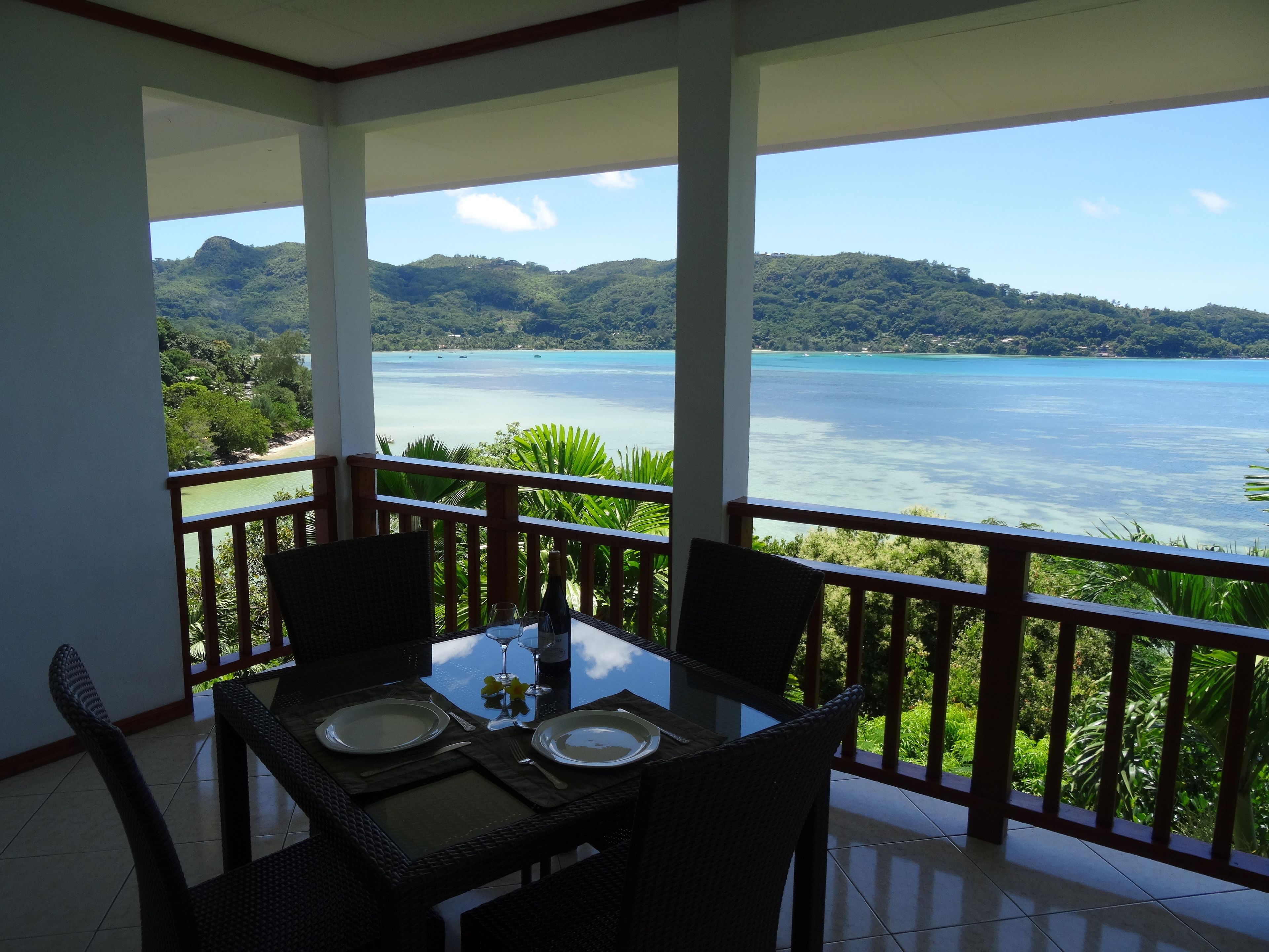 Un balcon avec une table dressée pour deux personnes, offrant une vue sur une mer turquoise et des collines verdoyantes.