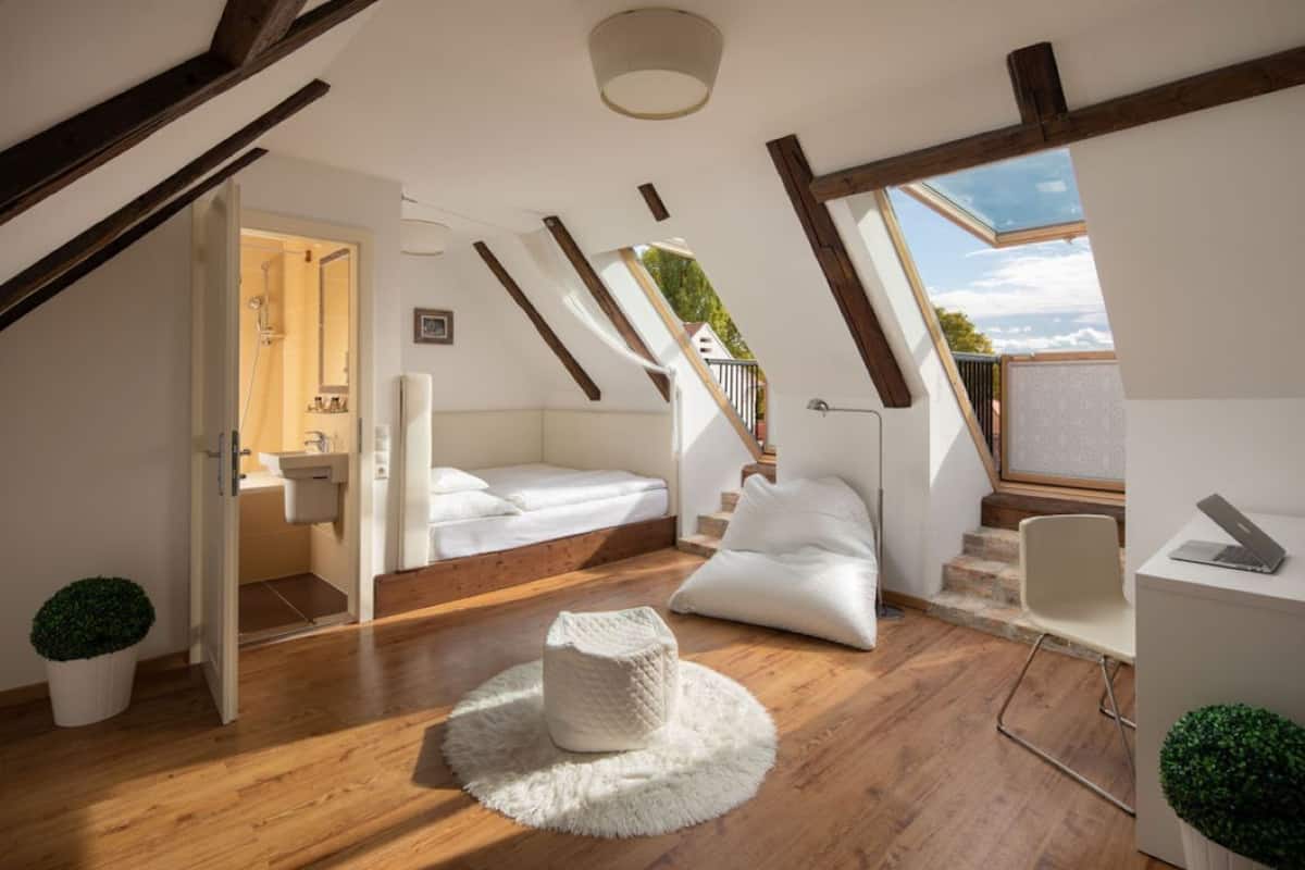 Un dormitorio tipo loft con piso de madera, una cama blanca, un escritorio con una computadora portátil y vistas al cielo a través de un tragaluz.