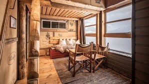 L’intérieur d’un chalet rustique, avec des murs en bois, une table en bois entourée de chaises, un banc agrémenté de coussins et une fenêtre donnant sur un paysage enneigé.