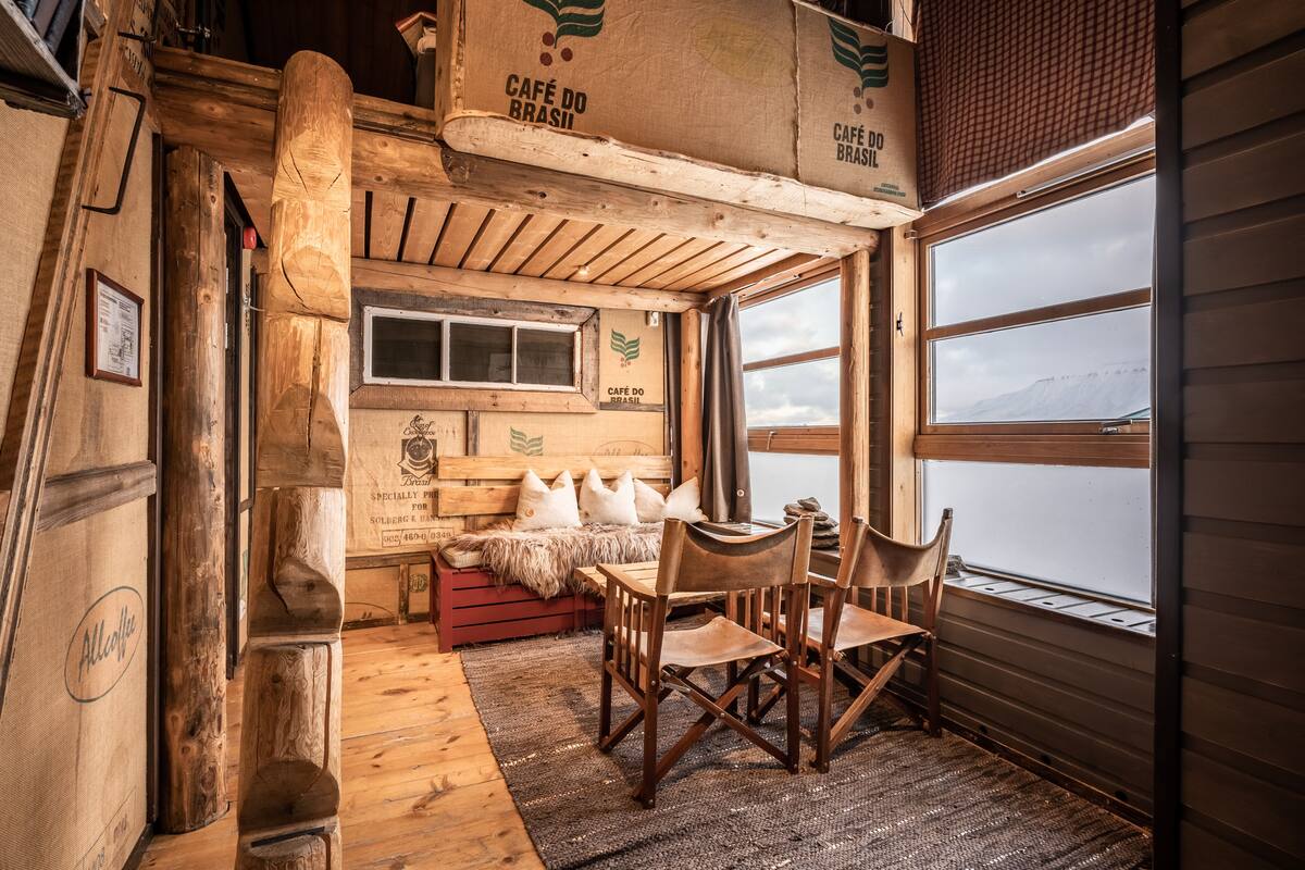 A rustic cabin interior with wooden walls, a wooden table with chairs, a bench with pillows, and a window overlooking a snowy landscape.