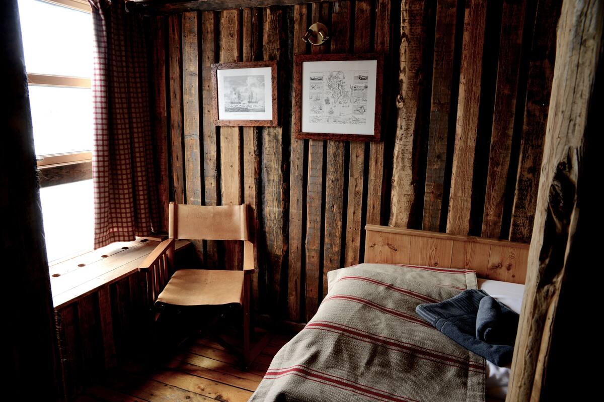 A rustic room with a wooden wall, a desk with a chair, and a bed with a striped blanket.