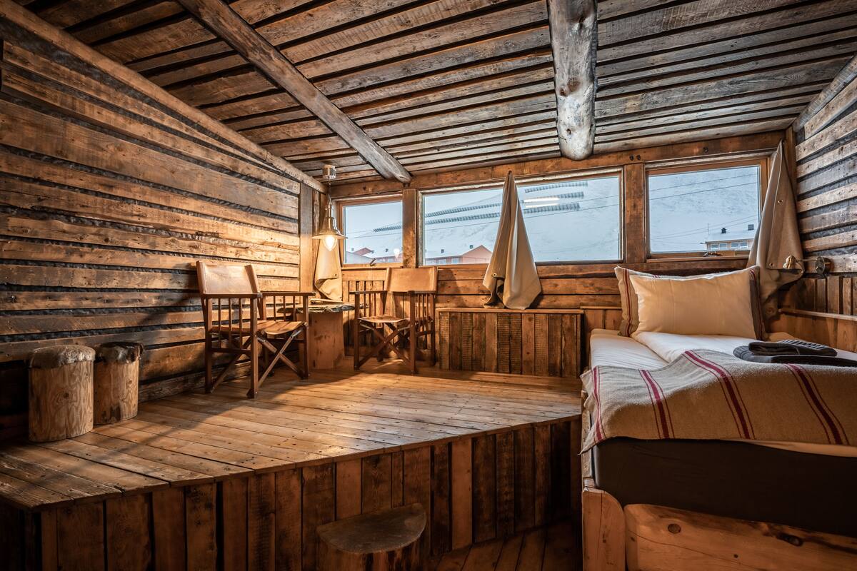 A cabin-style room with wooden walls and floors, a bed with a striped blanket, a desk with a lamp, and a window with a view of buildings.