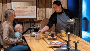 Une table en bois sur laquelle sont disposés divers aliments et boissons, un tableau encadré accroché au mur et une bibliothèque remplie de livres.