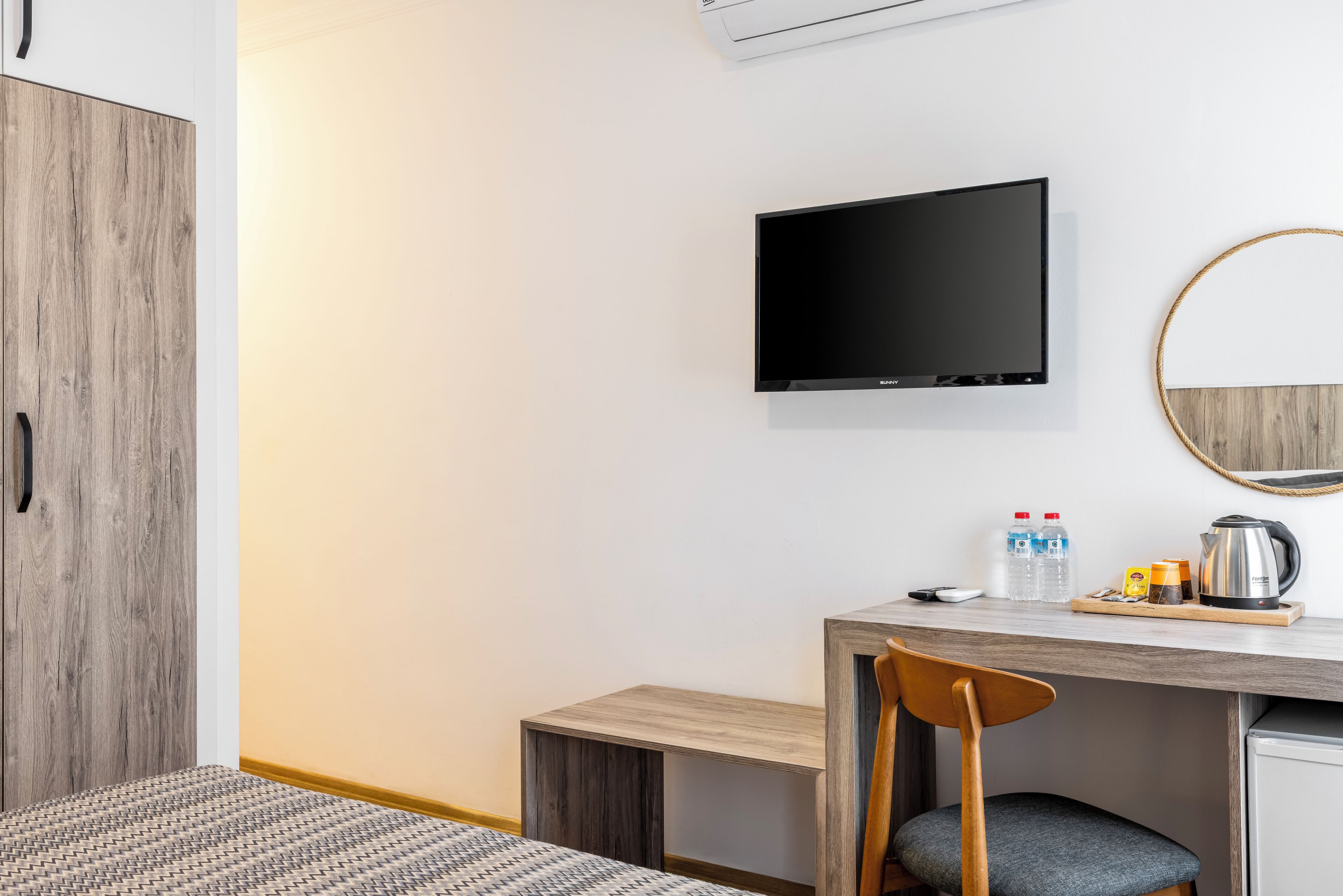 Ein modernes Hotelzimmer mit einem Holzschrank, einem flachbildfernseher an der Wand, einem Schreibtisch mit Stuhl und einer kleinen Küchenzeile.