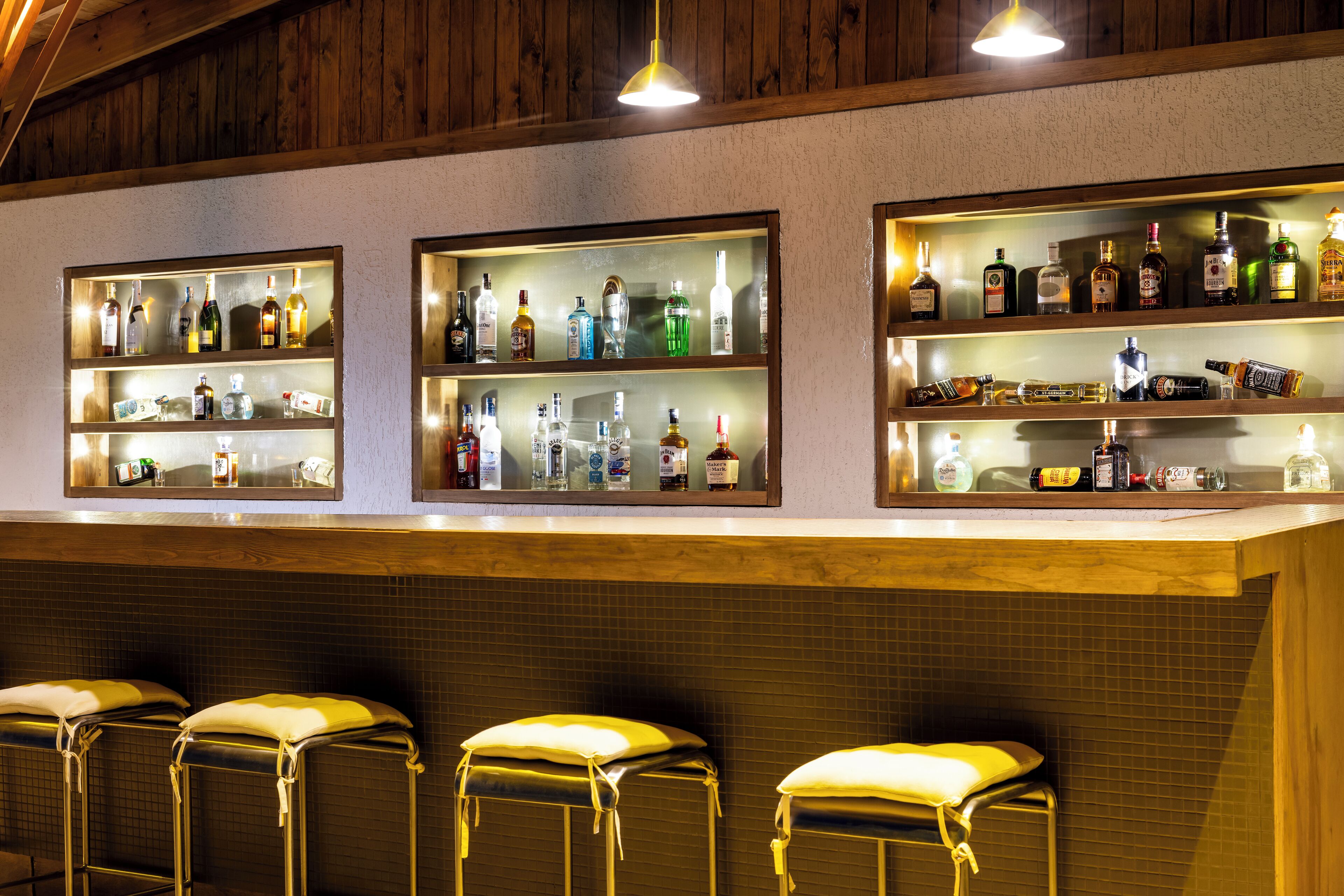 Un bar con varias estanterías llenas de botellas de licor, tres taburetes y paneles de madera en el techo.