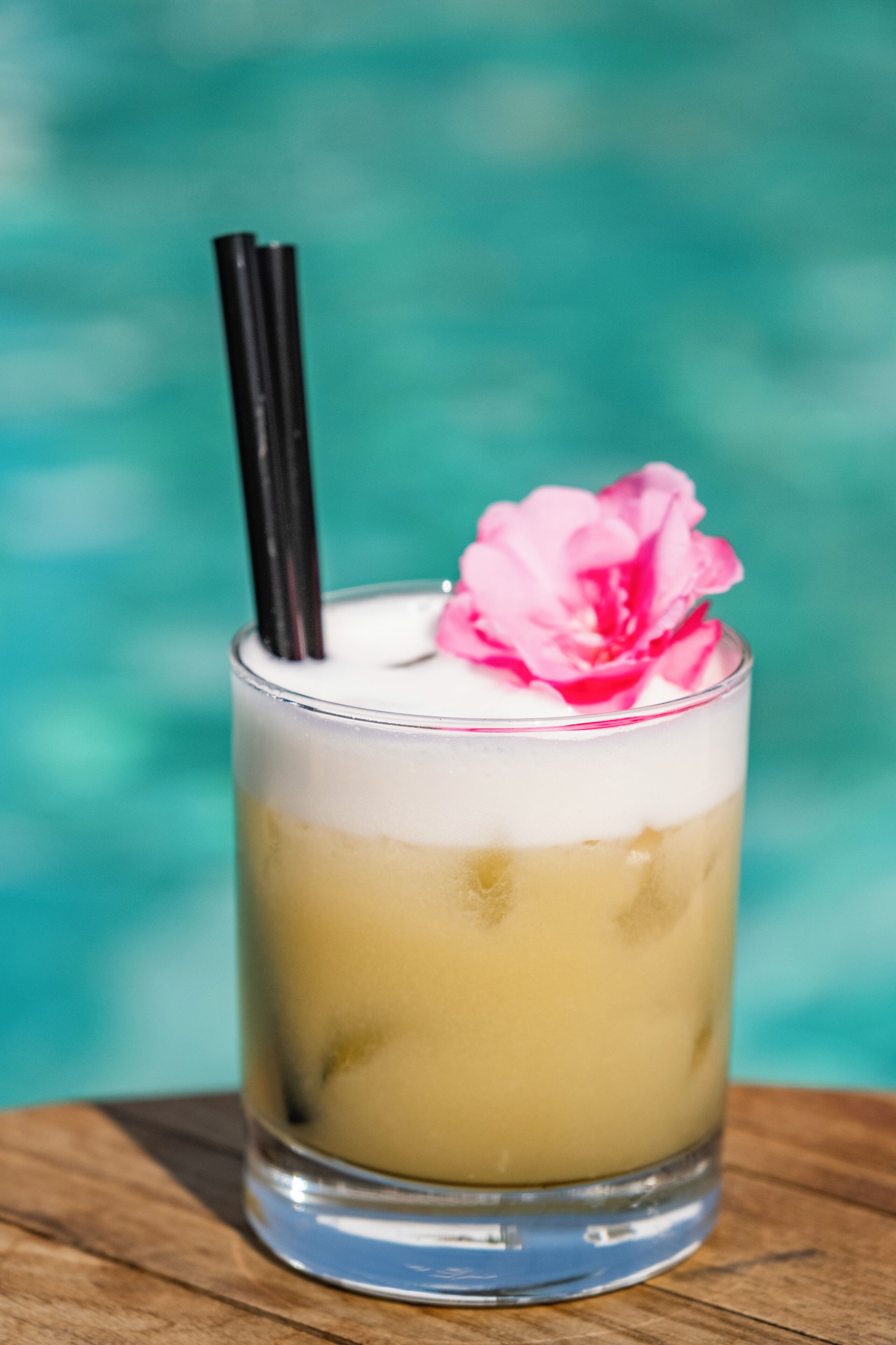 Een glas met gelaagde drankjes, een roze bloem als garnering en zwarte rietjes.