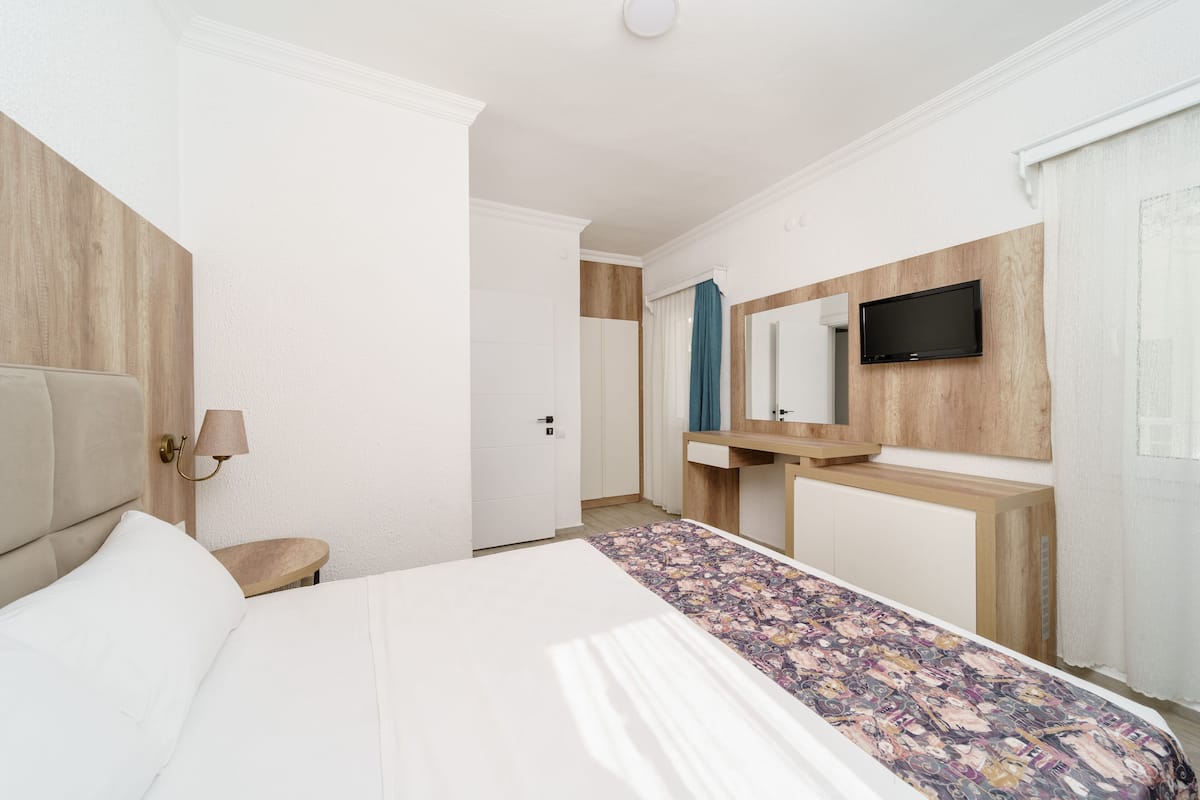 A hotel room with a bed, a bedside table with a lamp, a wooden headboard, a TV mounted on the wall, and a bathroom door.