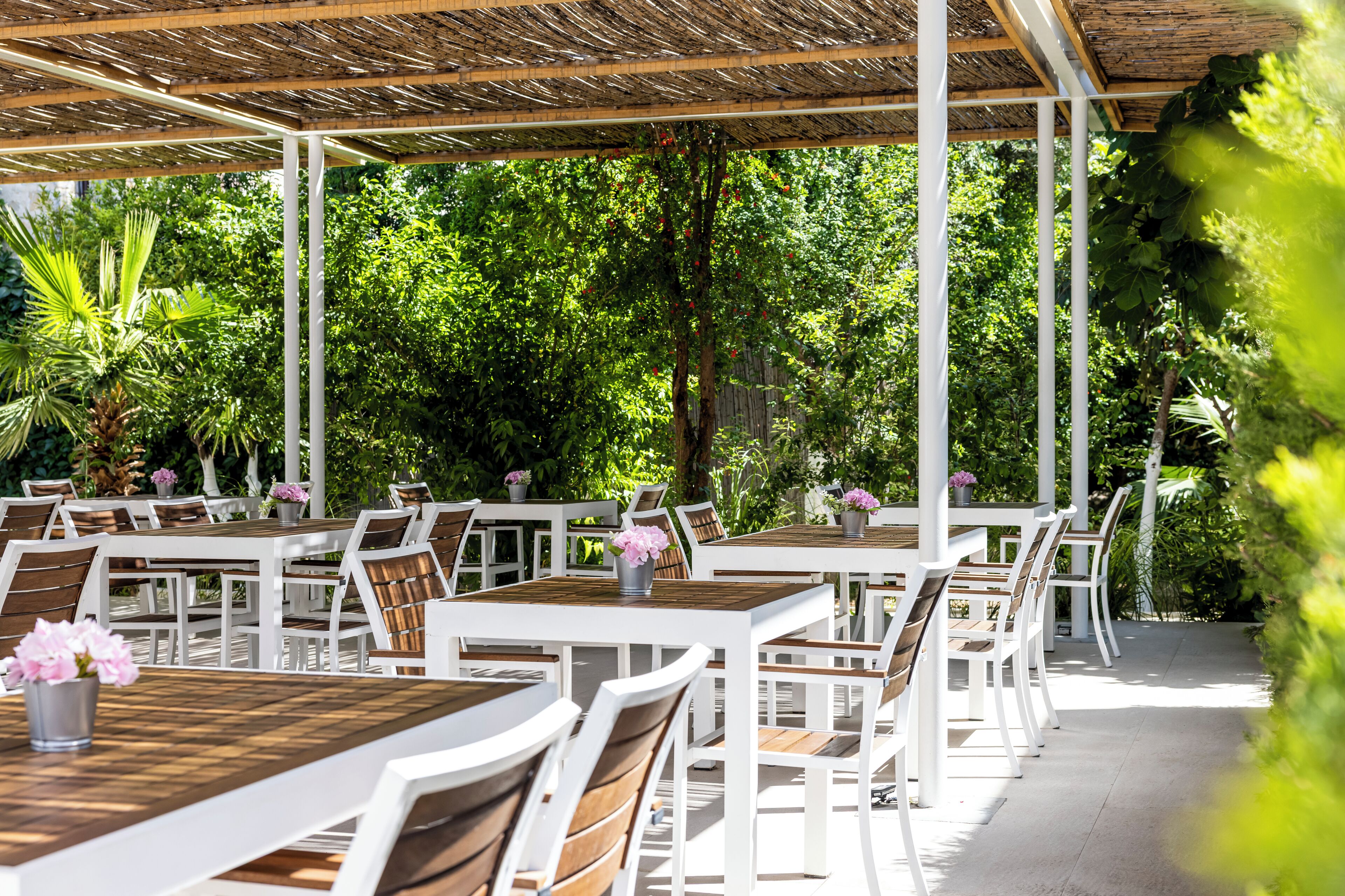An outdoor dining area with white tables and chairs, surrounded by lush greenery.