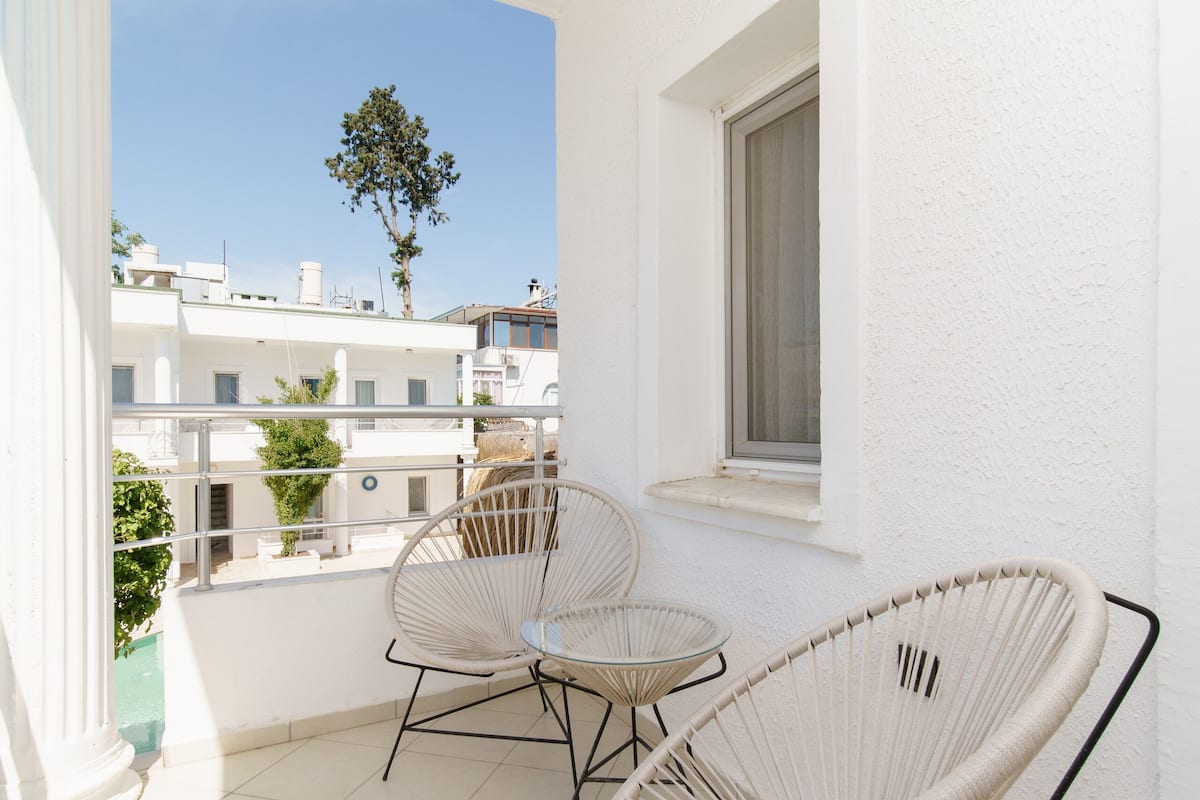 A balcony with two wicker chairs and a small table, white walls, and a window overlooking a residential area.