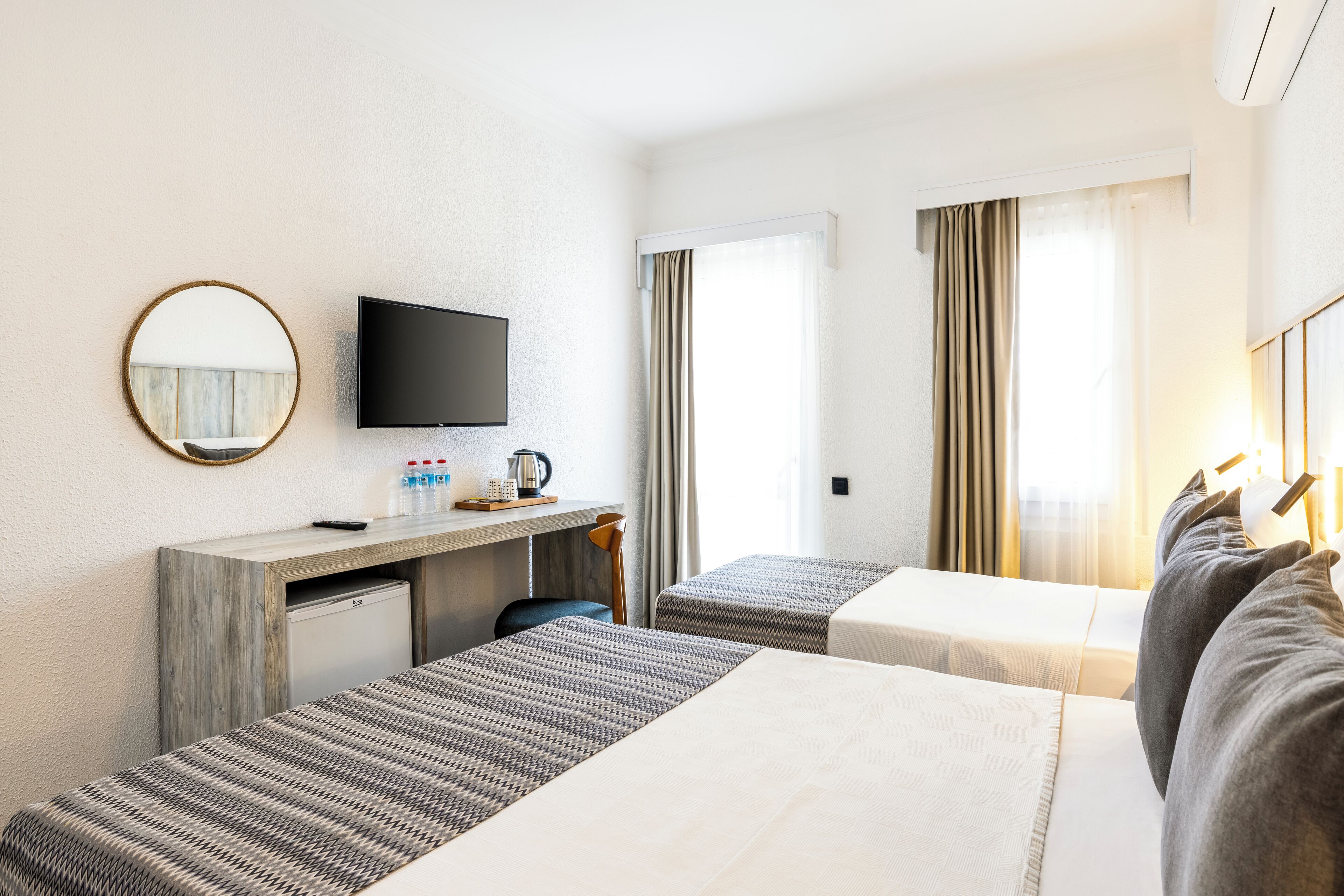 Habitación de hotel con dos camas, un escritorio con televisor, un refrigerador pequeño y una ventana con cortinas.