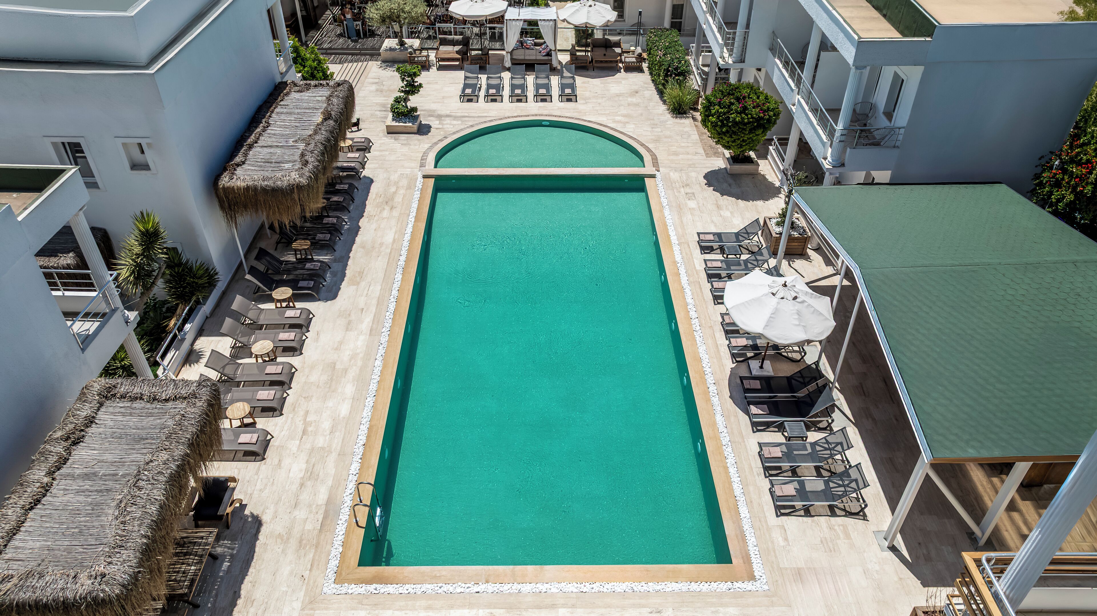 Una piscina rectangular con una estructura de techo de paja en un lateral, rodeada de sillas de descanso y sombrillas.