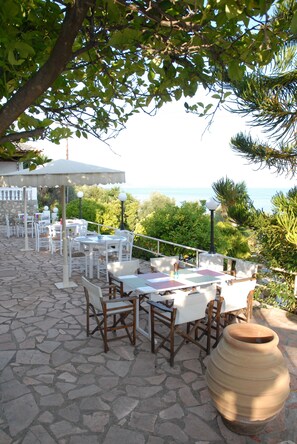 Una terrazza esterna con pavimento in pietra, tavoli e sedie bianche, immersa nel verde e con vista su un corpo d'acqua.