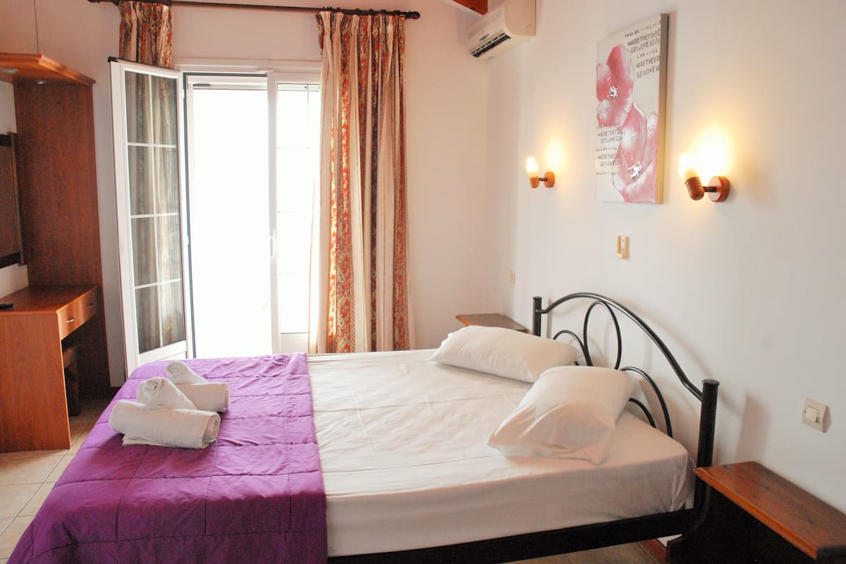A bedroom with a bed, pillows, a purple bedspread, a wooden desk, a chair, a window with curtains, and a wall-mounted light fixture.
