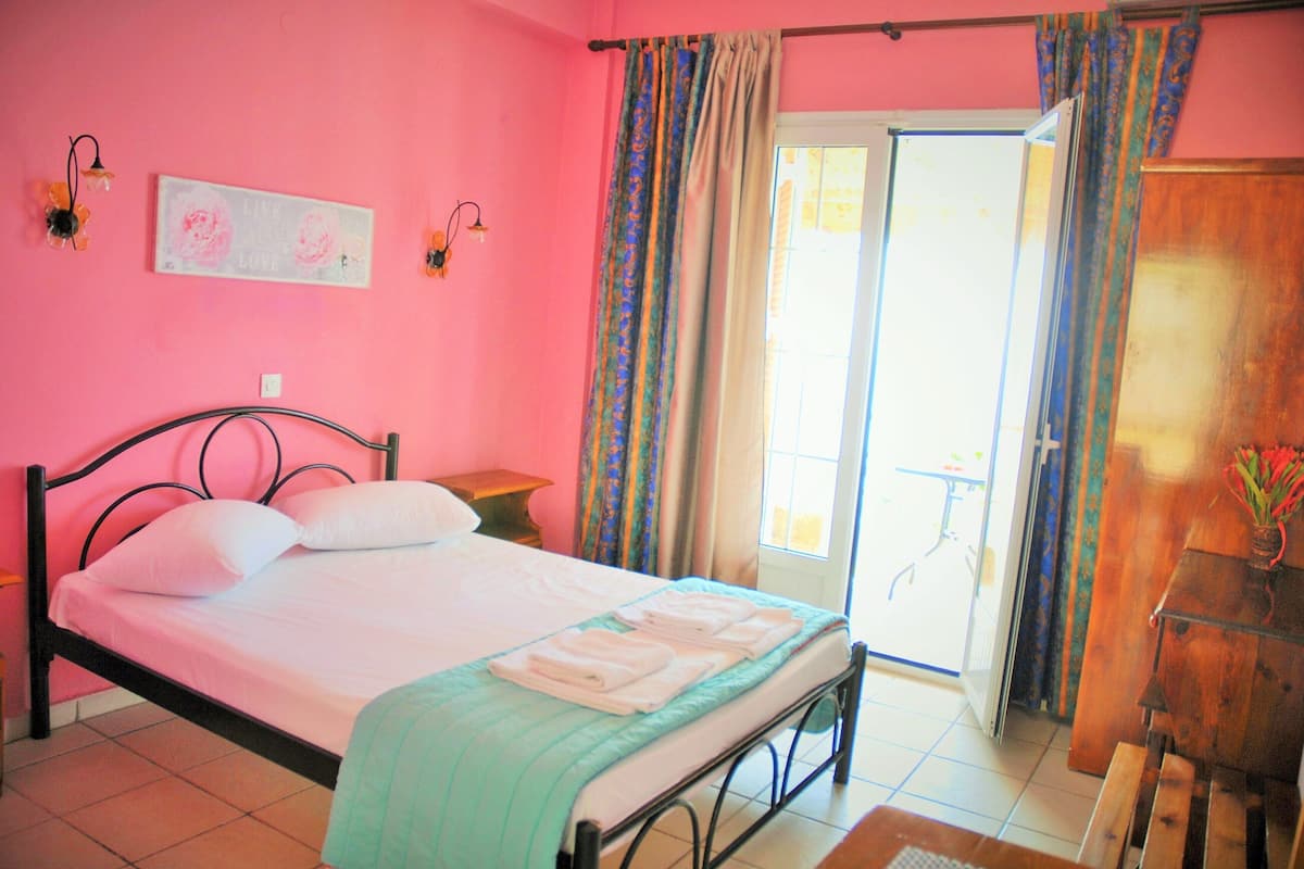 A bedroom with a bed, a nightstand, a wooden dresser, a window with curtains, and a door leading to a balcony.