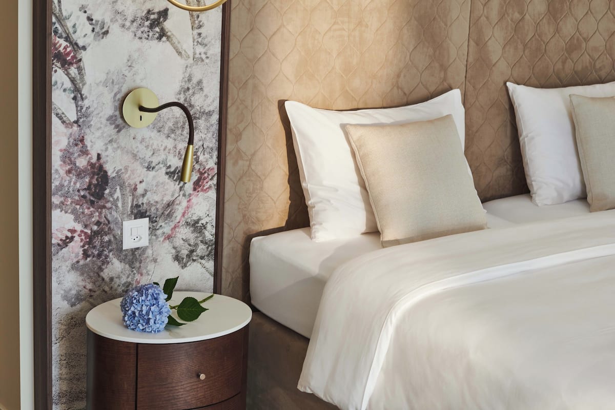 Una cama bien tendida con sábanas blancas y almohadas color beige, una mesita de noche redonda con un objeto decorativo azul, y una pared con textura y una lámpara fijada a la pared.