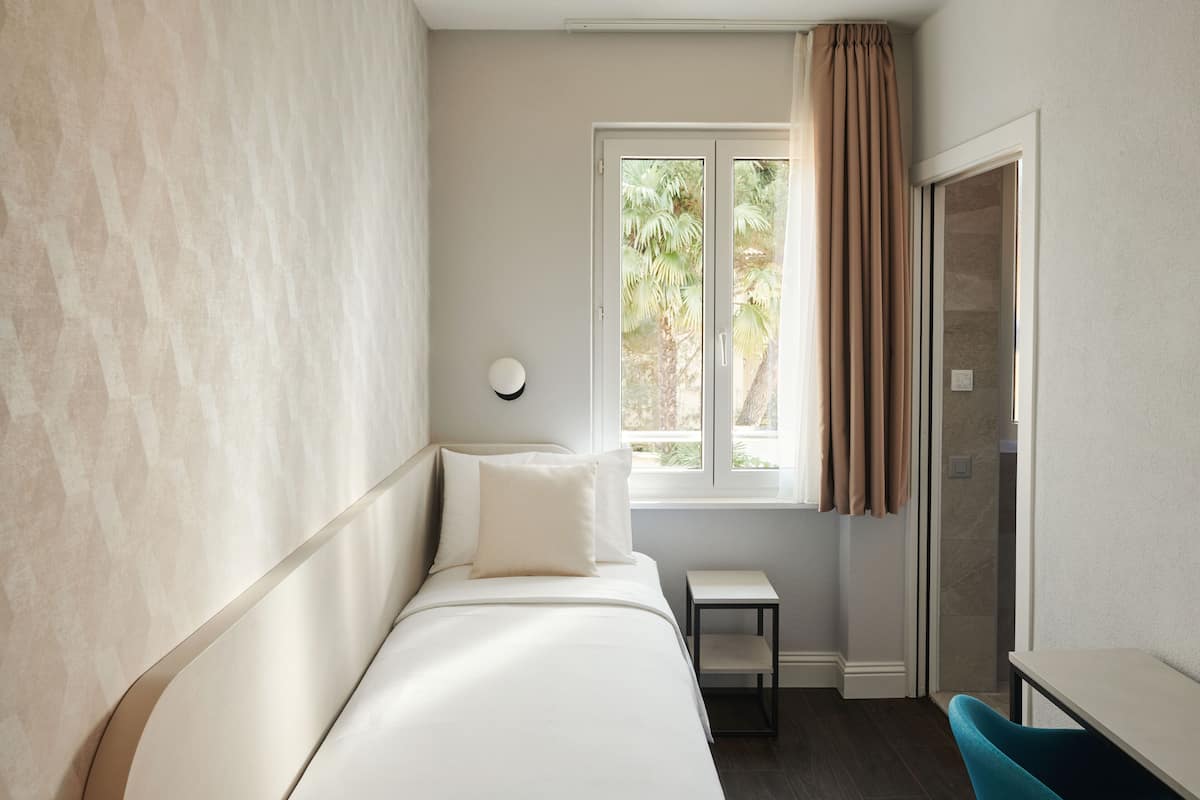 Un dormitorio con cama, una mesita de noche pequeña, una ventana con cortinas y una puerta.