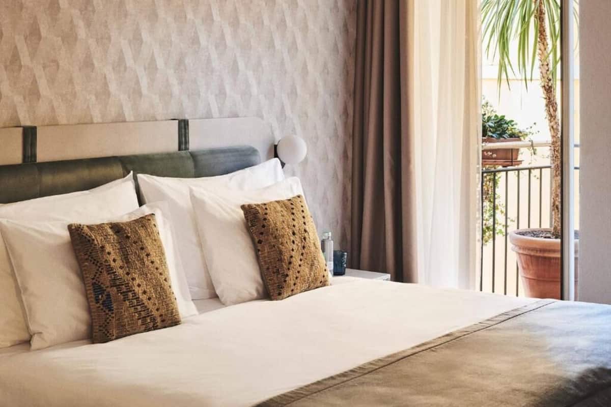 Una habitación de hotel con cama, cabecera y vistas al balcón con plantas en macetas.