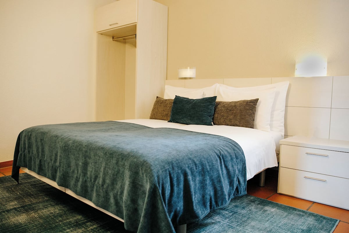 Una cama bien hecha con una colcha azul oscuro, almohadas blancas y una almohada decorativa verde. A la derecha de la cama, se encuentra una mesita de noche blanca con cajones.