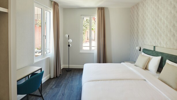 Habitación de hotel con cama, escritorio y silla. Cuenta con ventana con cortinas, lámpara de pie y vista al exterior.
