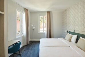 Habitación de hotel con cama, escritorio y silla. Cuenta con ventana con cortinas, lámpara de pie y vista al exterior.