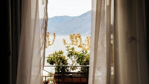 Vista a través de cortinas traslúcidas de un balcón con planta en maceta y barandilla, con vista a una masa de agua y montañas distantes.