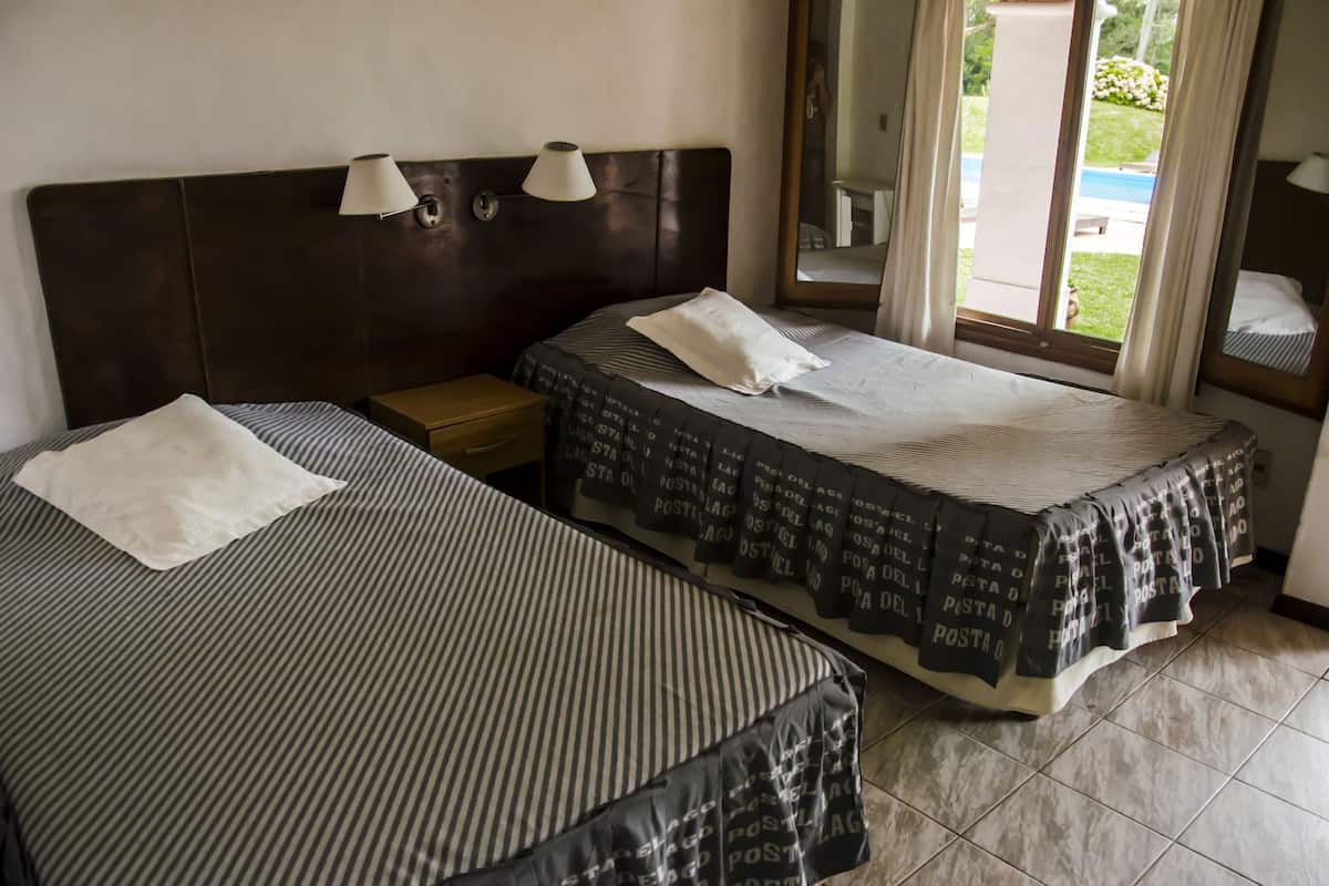 Habitación de hotel con dos camas, cada una con almohada y cabecera. Hay dos lámparas de noche y una ventana con cortinas.