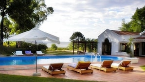 Piscina com espreguiçadeiras e guarda-sóis, cercada por um gramado bem cuidado e um edifício com telhado em azulejos.