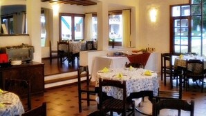 Uma sala de jantar com mesas postas, vigas de madeira no teto e grandes janelas que permitem a entrada de luz natural.