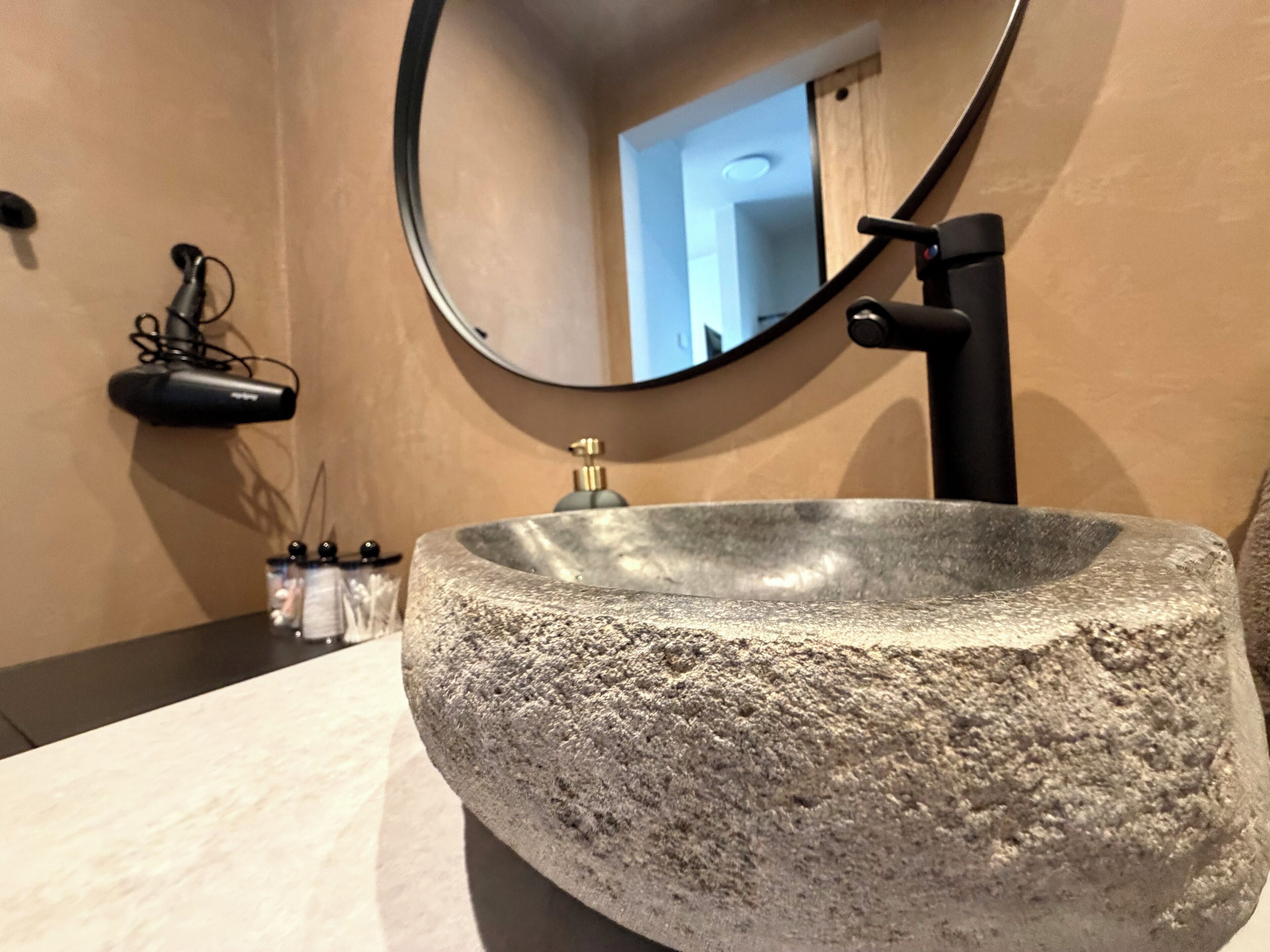 Une salle de bain avec un lavabo en pierre, un miroir rond et un robinet noir.