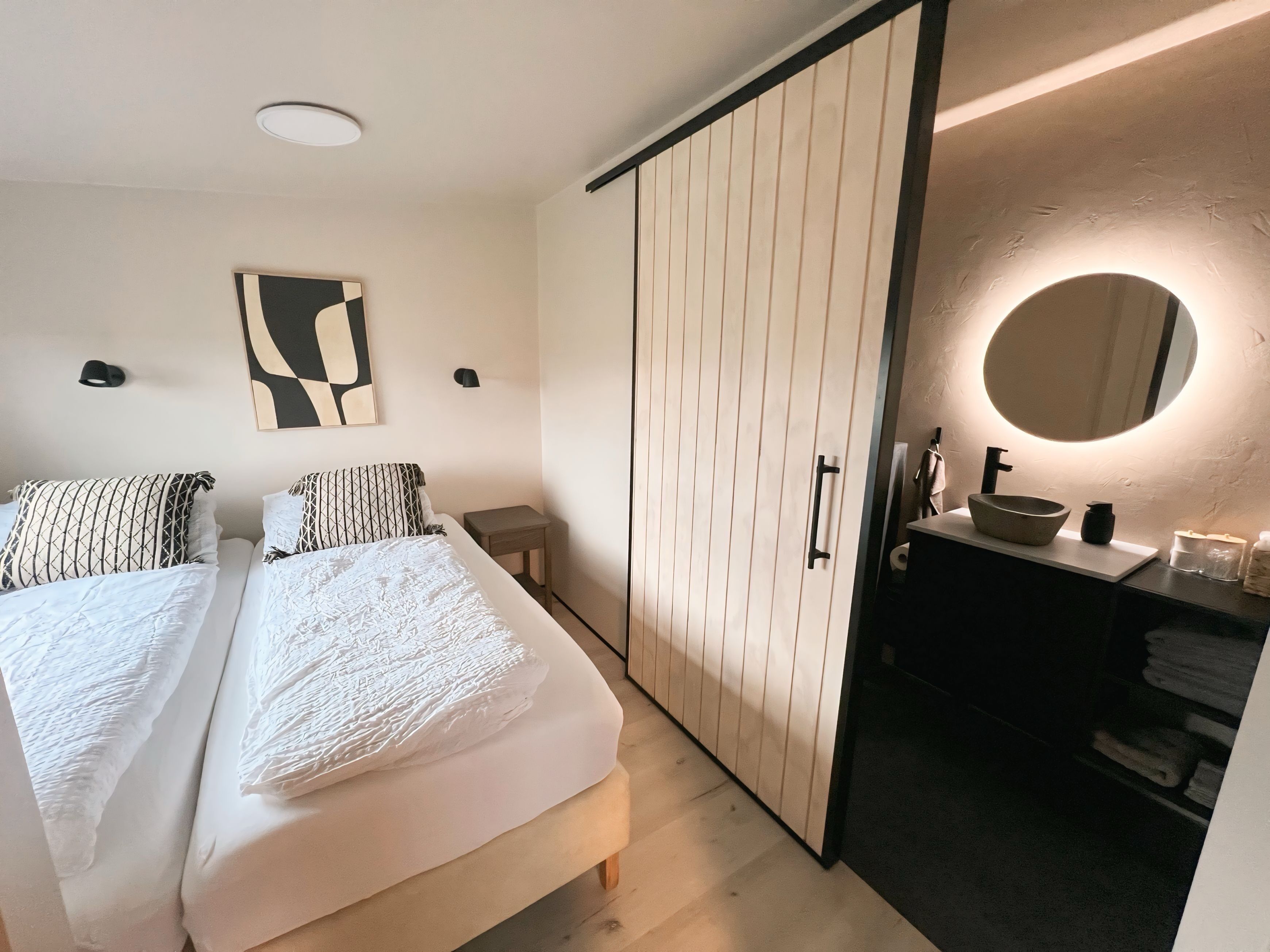 A modern bedroom with a bed, bedside tables, a wardrobe, a bathroom with a sink and mirror.