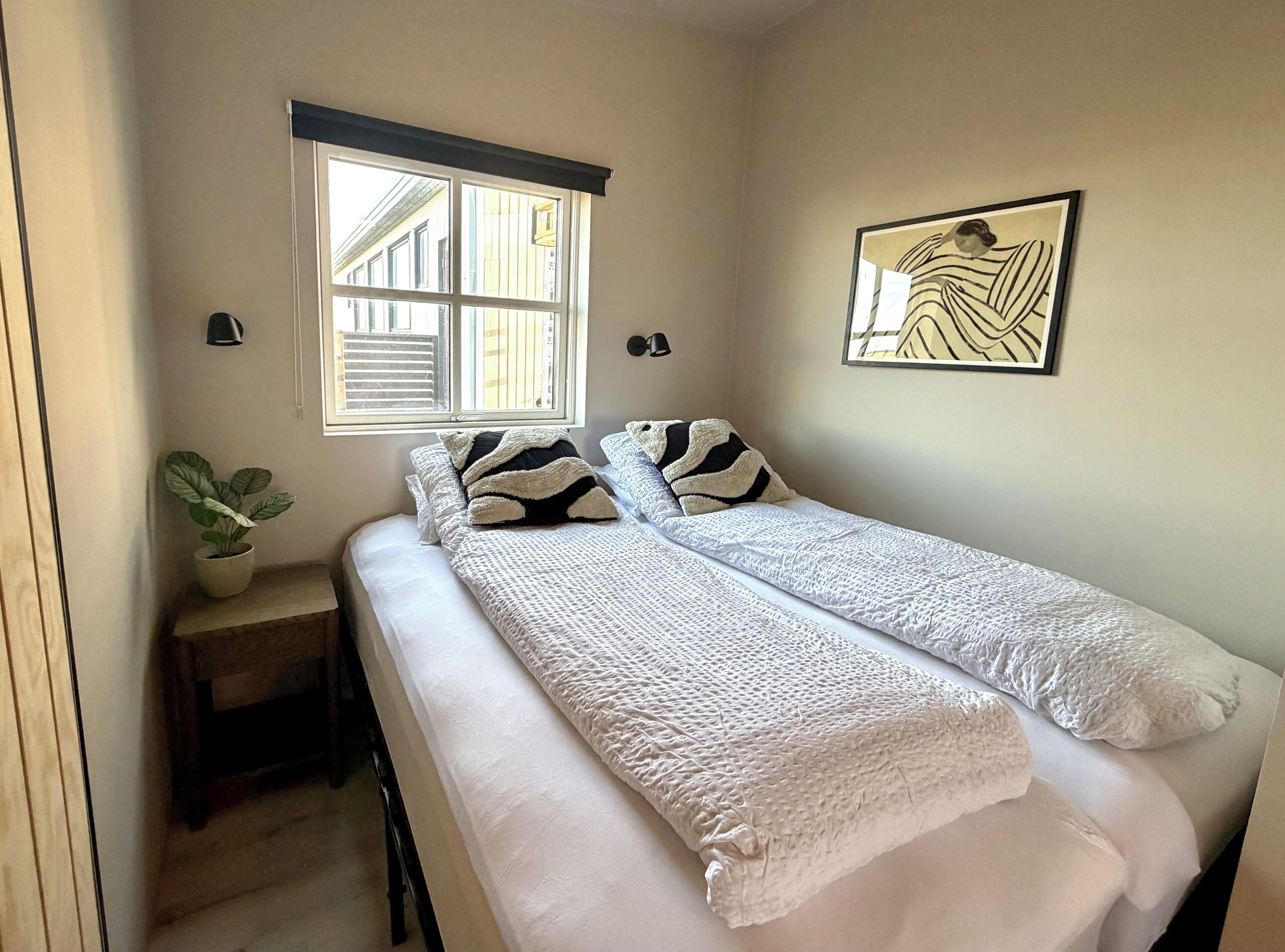 Une chambre avec deux lits, une fenêtre avec des stores, une plante en pot et un tableau encadré au mur.