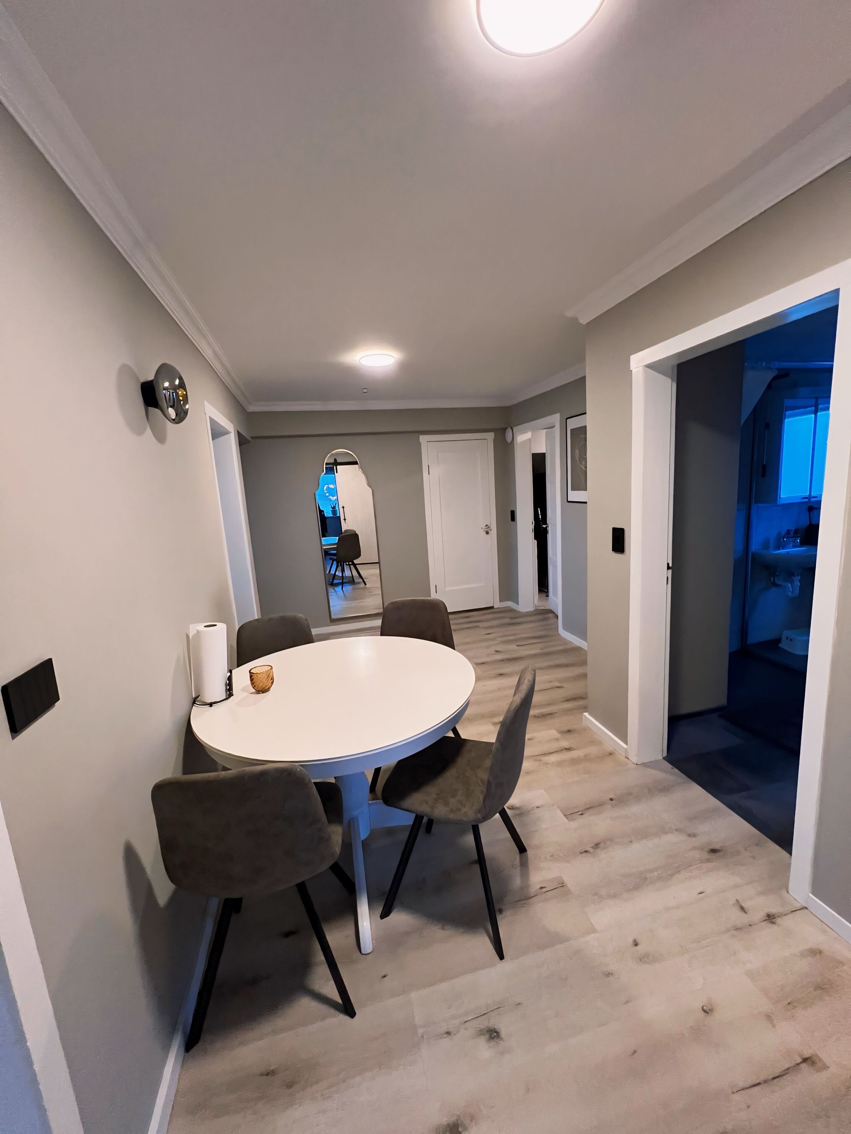 Un comedor moderno con una mesa redonda y cuatro sillas, un aplique de pared y un pasillo que conduce a otras habitaciones.